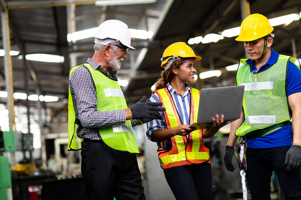 Three industrial workers wearing safety vests and hard hats standing inside a factory, gathered around a laptop held by the woman in the center, discussing or reviewing something together.
