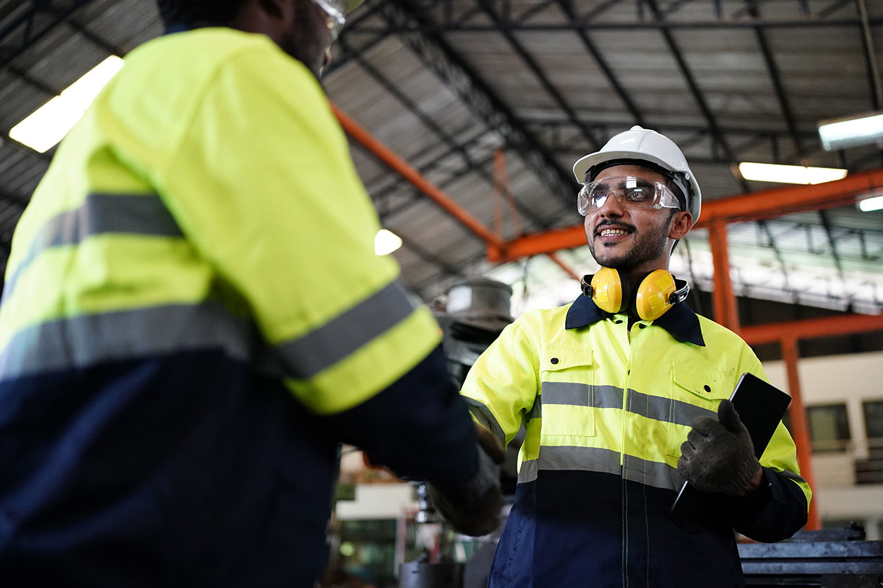 Two industrial workers in high-visibility jackets and hard hats shaking hands inside a factory, with one worker smiling while holding a tablet and wearing protective goggles and ear protection around his neck.