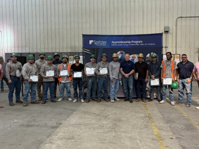 Group photo of Spitzer Apprenticeship Program participants, graduates, and company leaders holding completion certificates at a manufacturing facility.